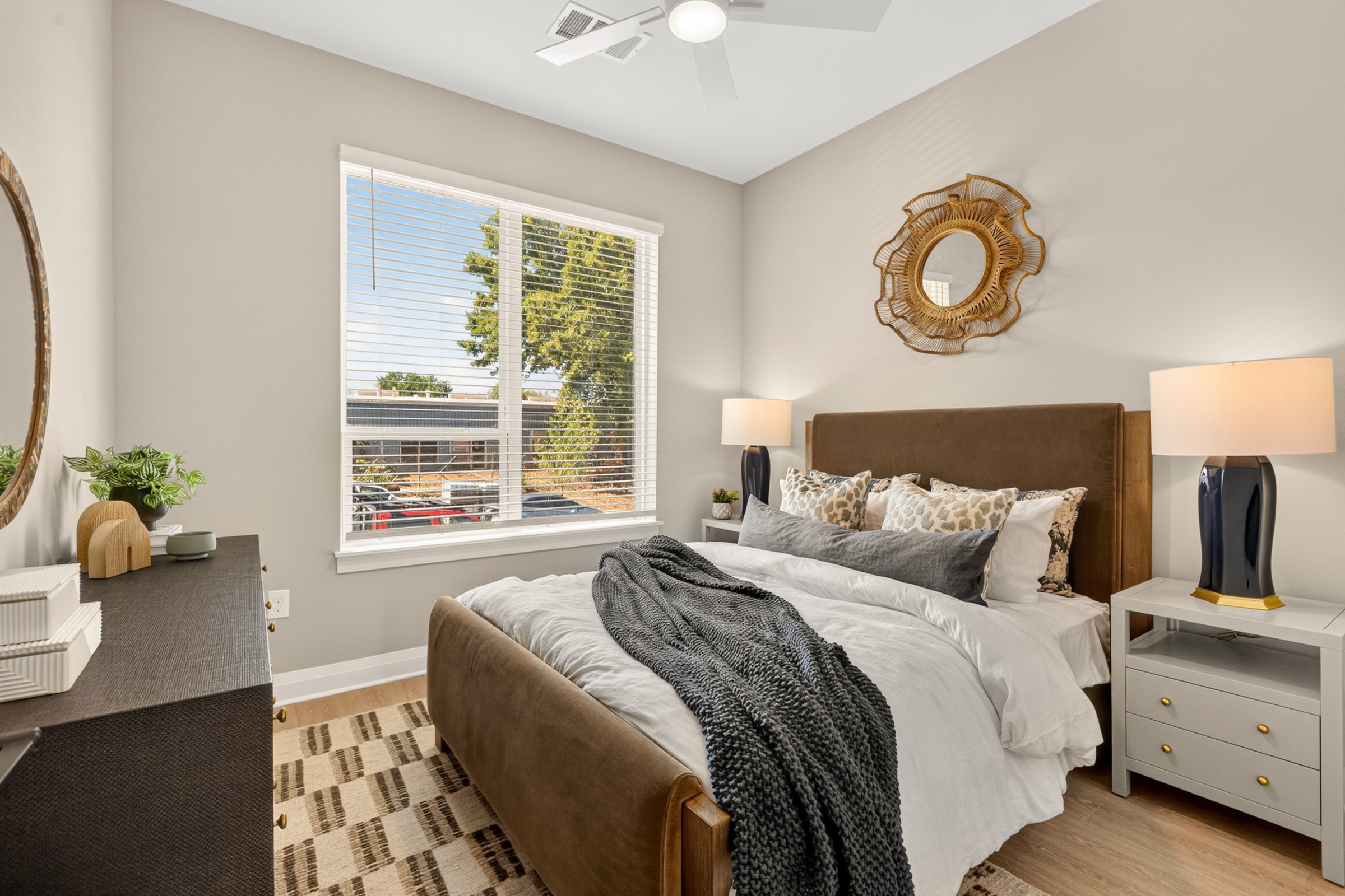 Bright bedroom with a large window, brown bed, neutral decor, and a modern round mirror above the headboard.