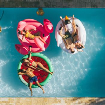 Three people relaxing on colorful pool floats—a flamingo, a unicorn, and a watermelon—seen from above.