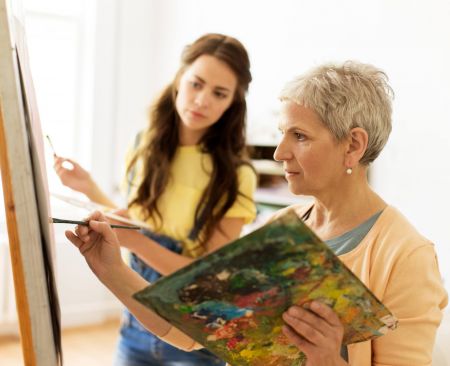 Two women painting together; one holds a palette while the other paints on a canvas in a bright room.