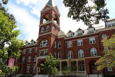 Red brick building with a clock tower, arched windows, and a tree in the foreground under a blue sky.